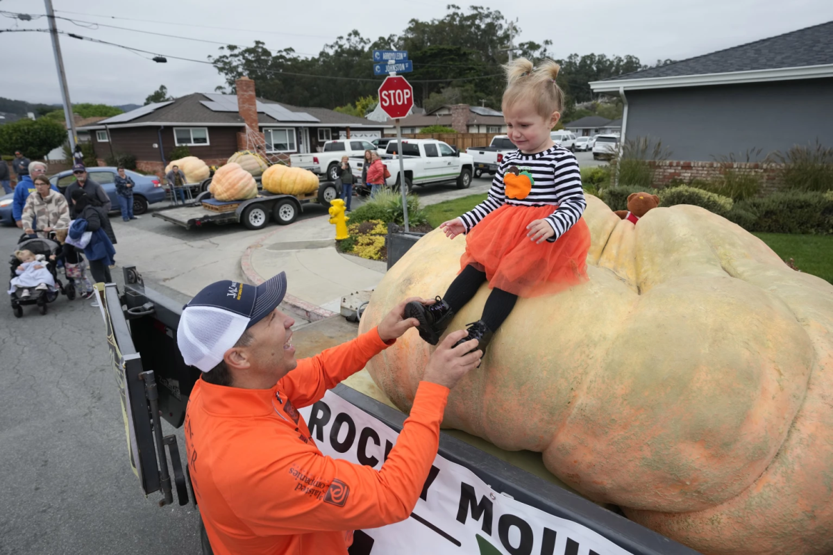 A little girl in an orange skirt and striped dress sits on top of a pumpkin. A man in an orange long sleeve shirt holds her feet steady.