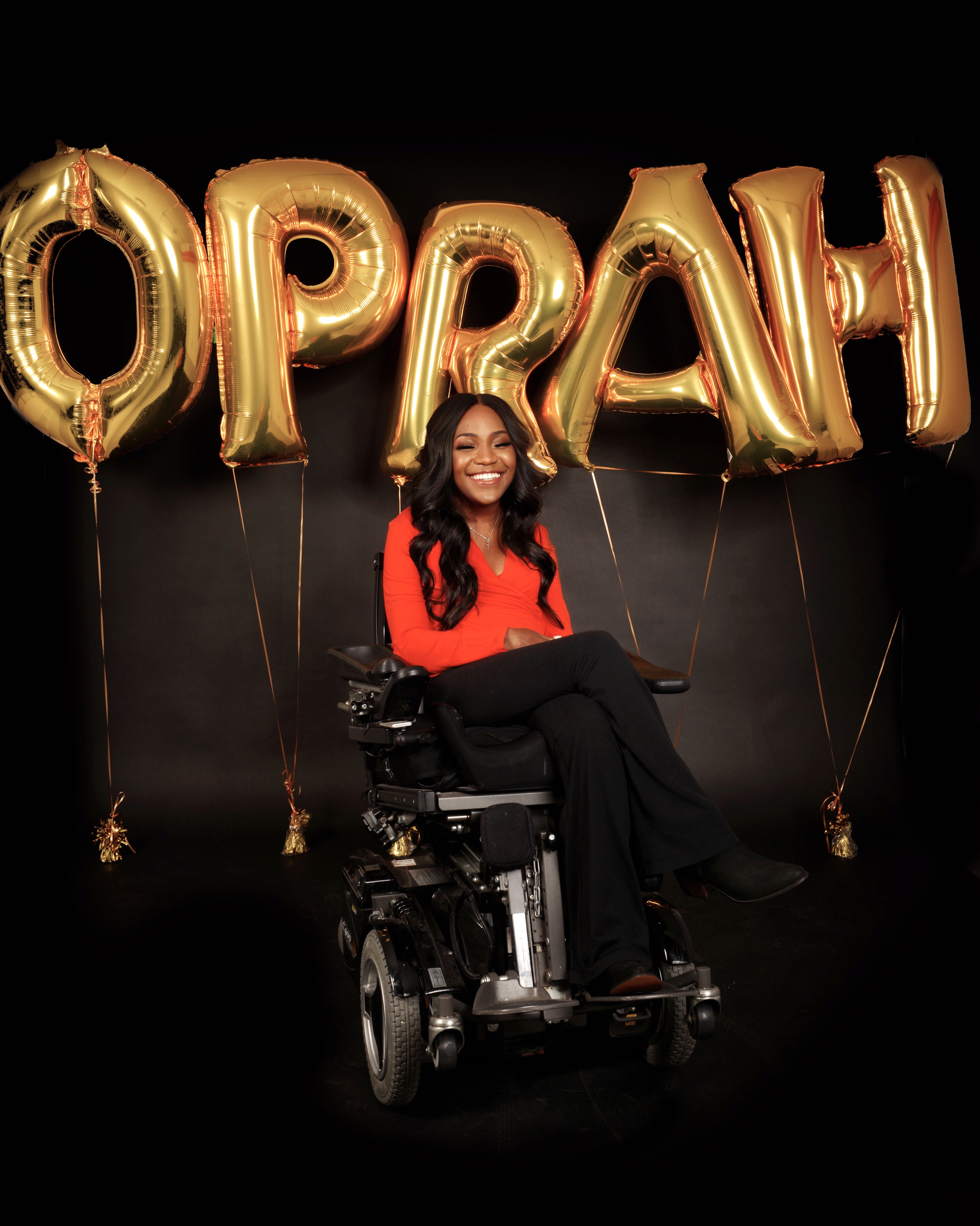 A smiling woman in a wheelchair with balloons spelling Oprah behind her