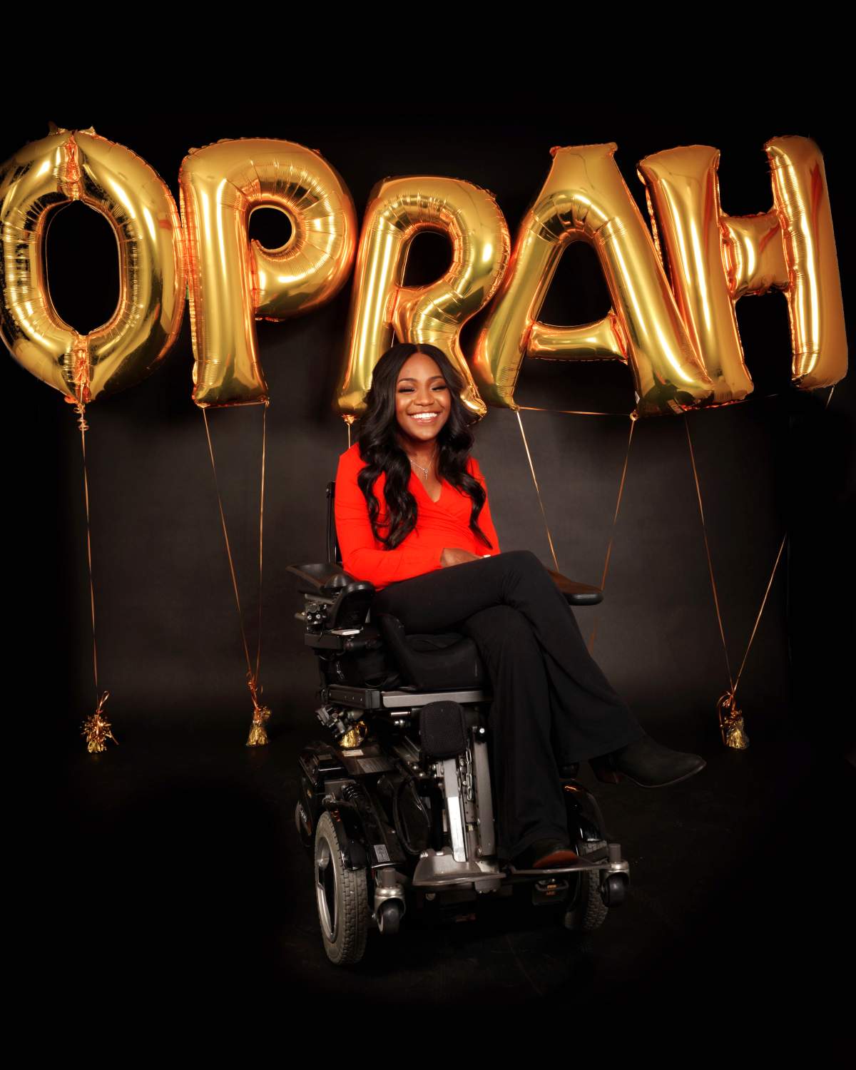 A smiling woman in a wheelchair with balloons spelling Oprah behind her