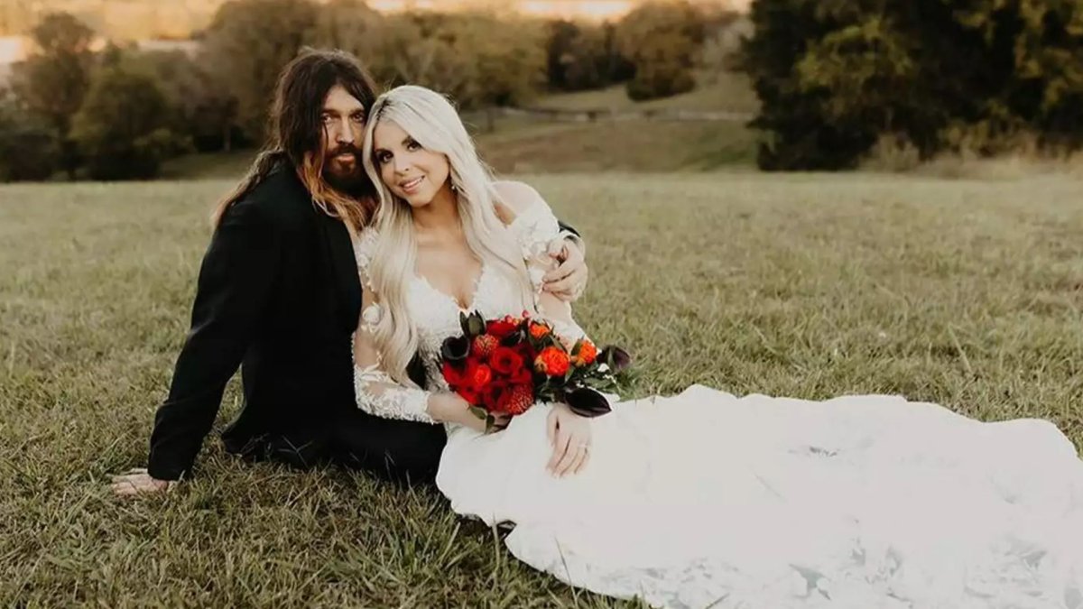 Billy Ray Cyrus and Firerose sit on a grassy hill in their wedding garb,