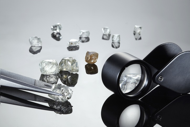 Uncut diamonds on a black perspex background with reflections, tweezers and a loupe.