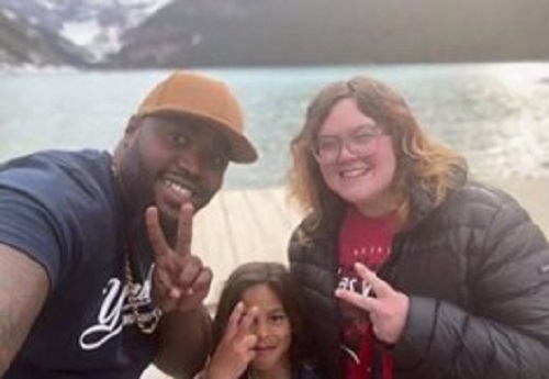 A man poses with his wife and son in front of a lake. They look into the camera, smiling, and make peace signs.