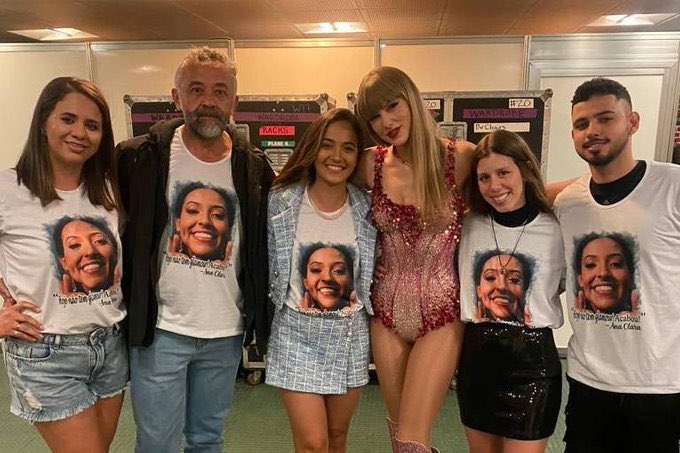 Taylor Swift with the family of Ana Clara Benevides Machado. Swift is wearing a pink bodysuit. The family members are wearing white T-shirts with Machado's face printed on them.