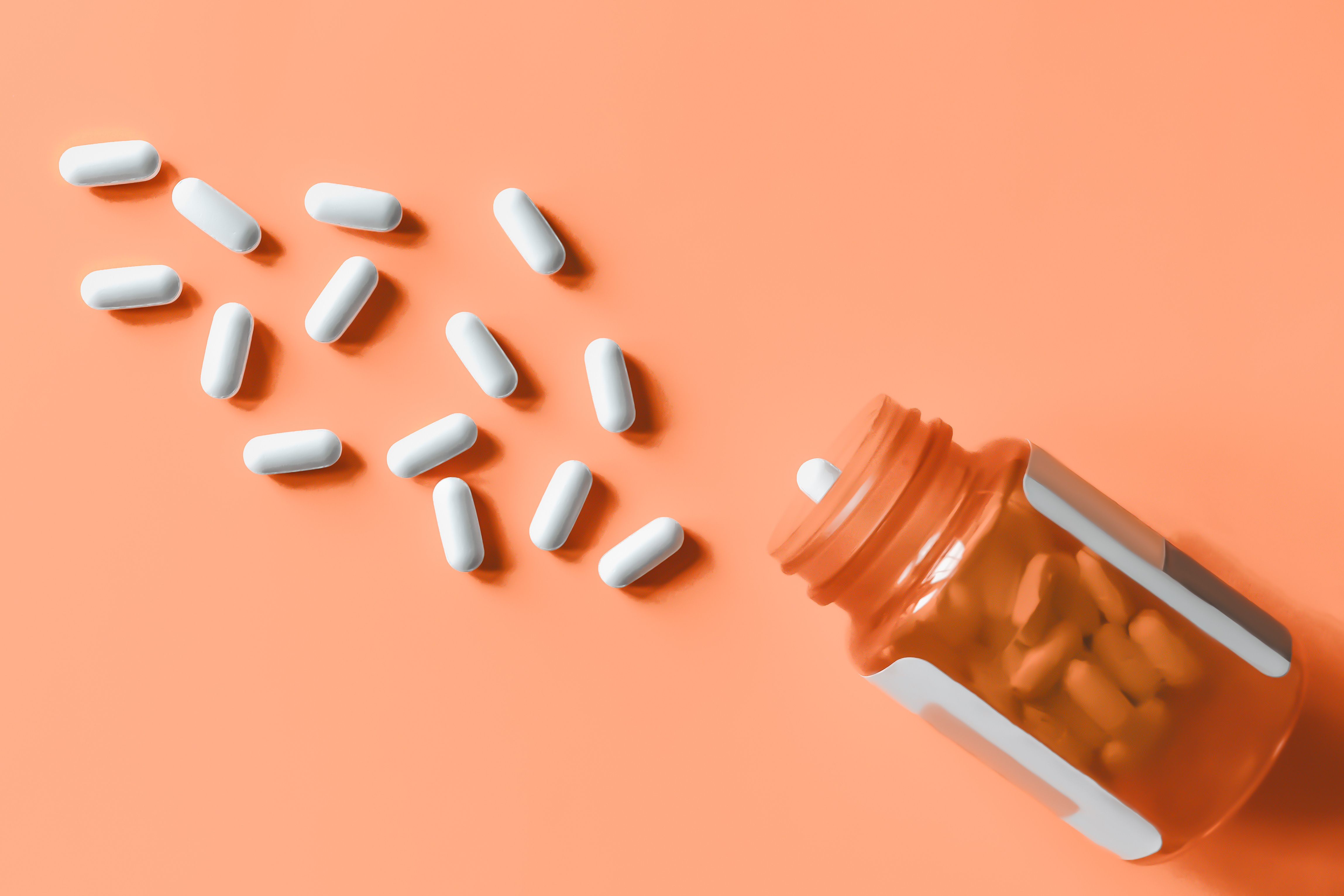 White pills spilling out of prescription bottle onto orange surface.