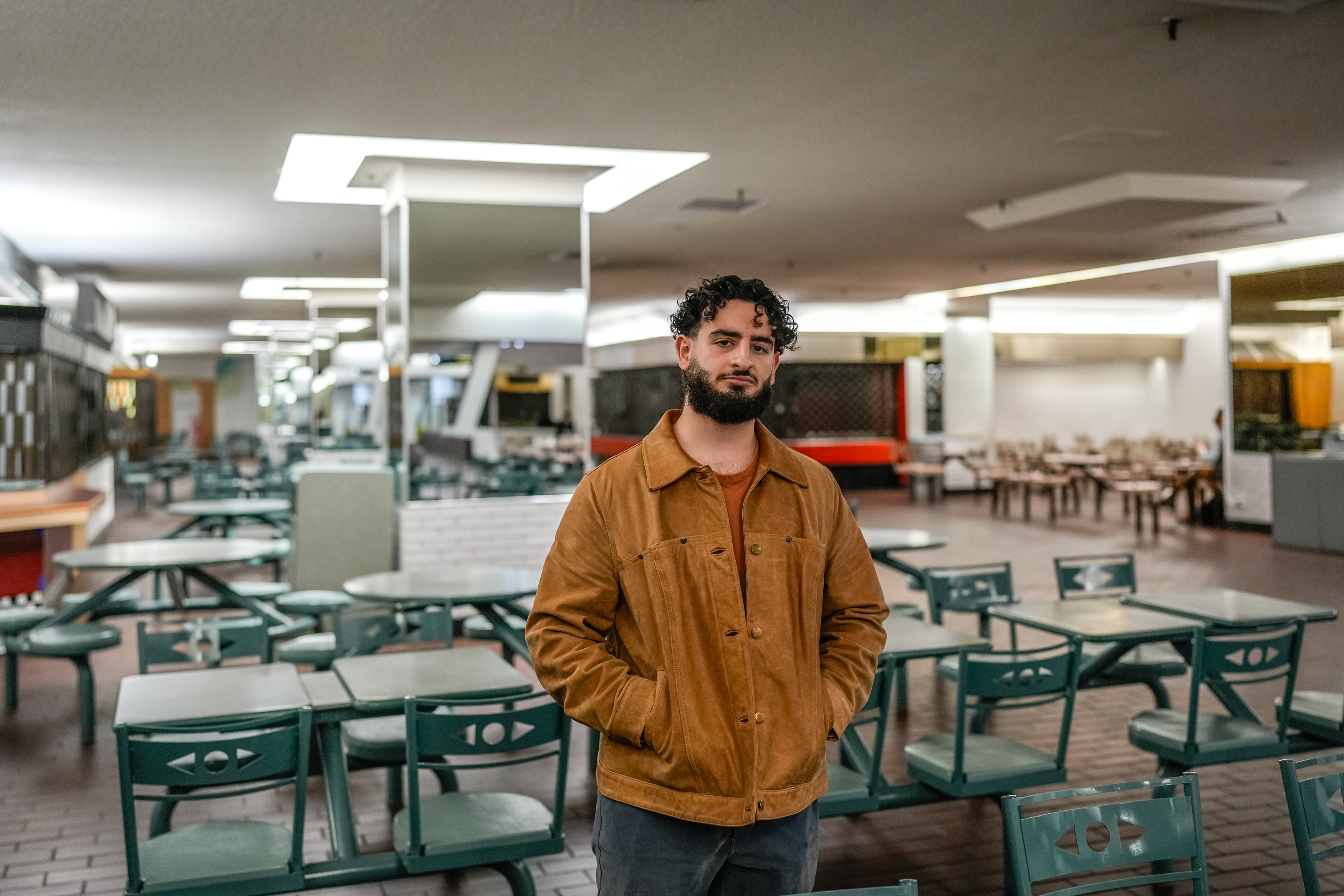 Aryeh Bookbinder wears a yellow-brown jacket and stands in the middle of an empty food court.
