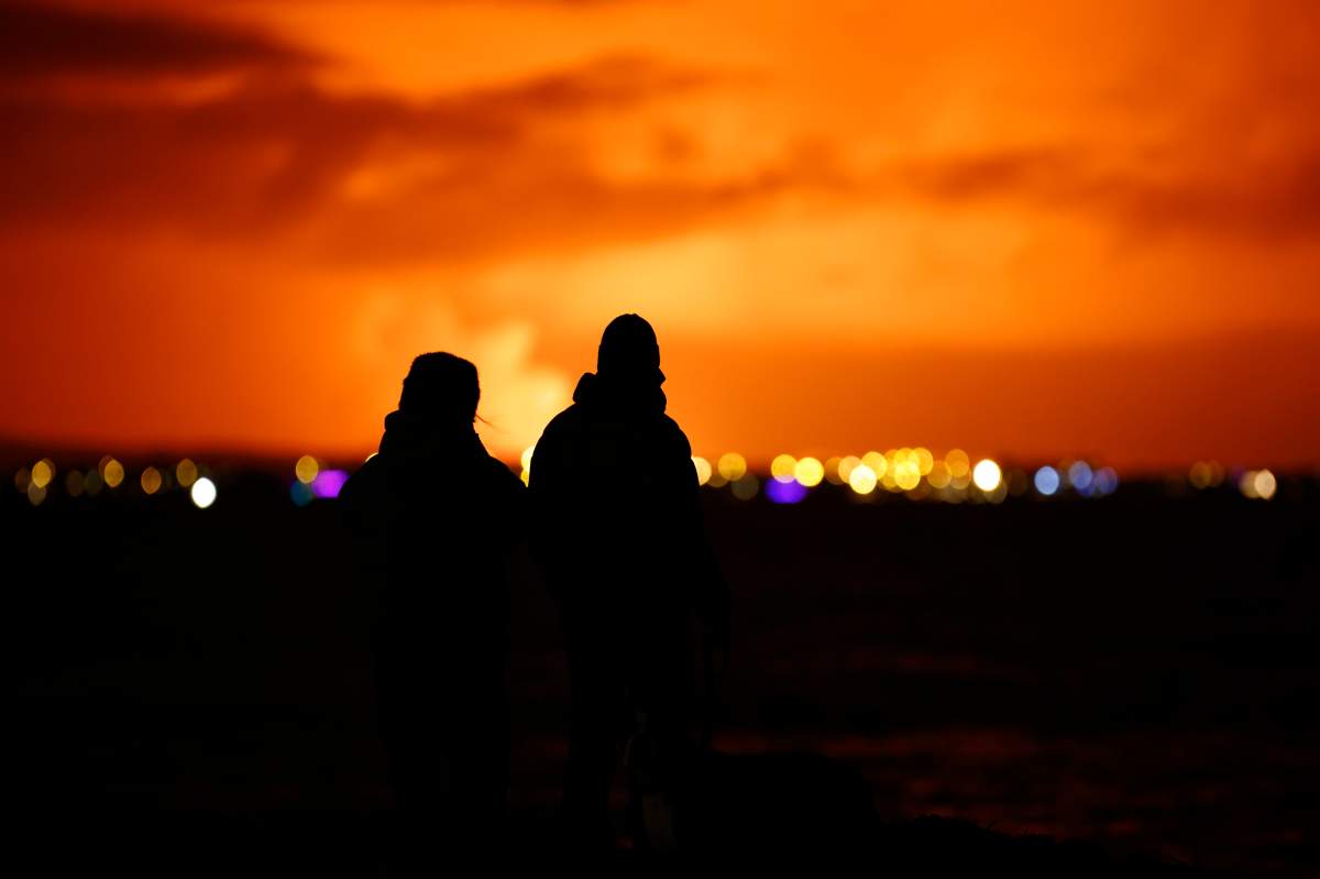 People watch as the night sky is illuminated caused by the eruption of a volcano on the Reykjanes peninsula of south-west Iceland seen from the capital city of Reykjavik, Monday Dec. 18, 2023. (AP Photo/Brynjar Gunnarsson)