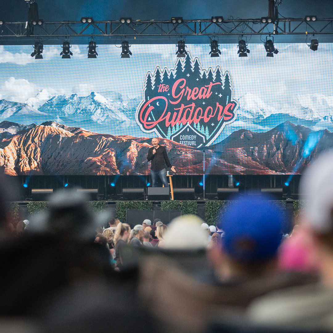 A comedian on an outdoor stage with a backdrop photo of mountains.