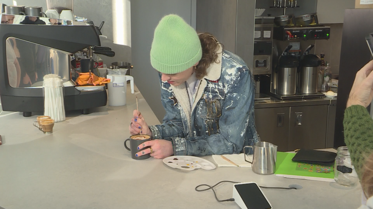 A man with a green hat and a leather jacket paints a drink with a wooden tool.