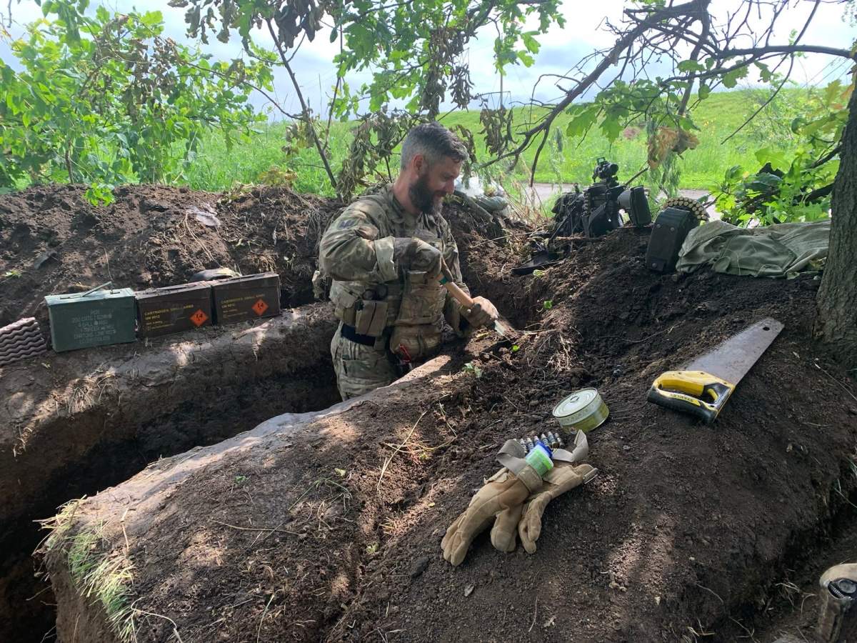 A soldier in fatigues digs a trench with a short shovel. He is waist deep in the trench, which is surrounded by green foliage.