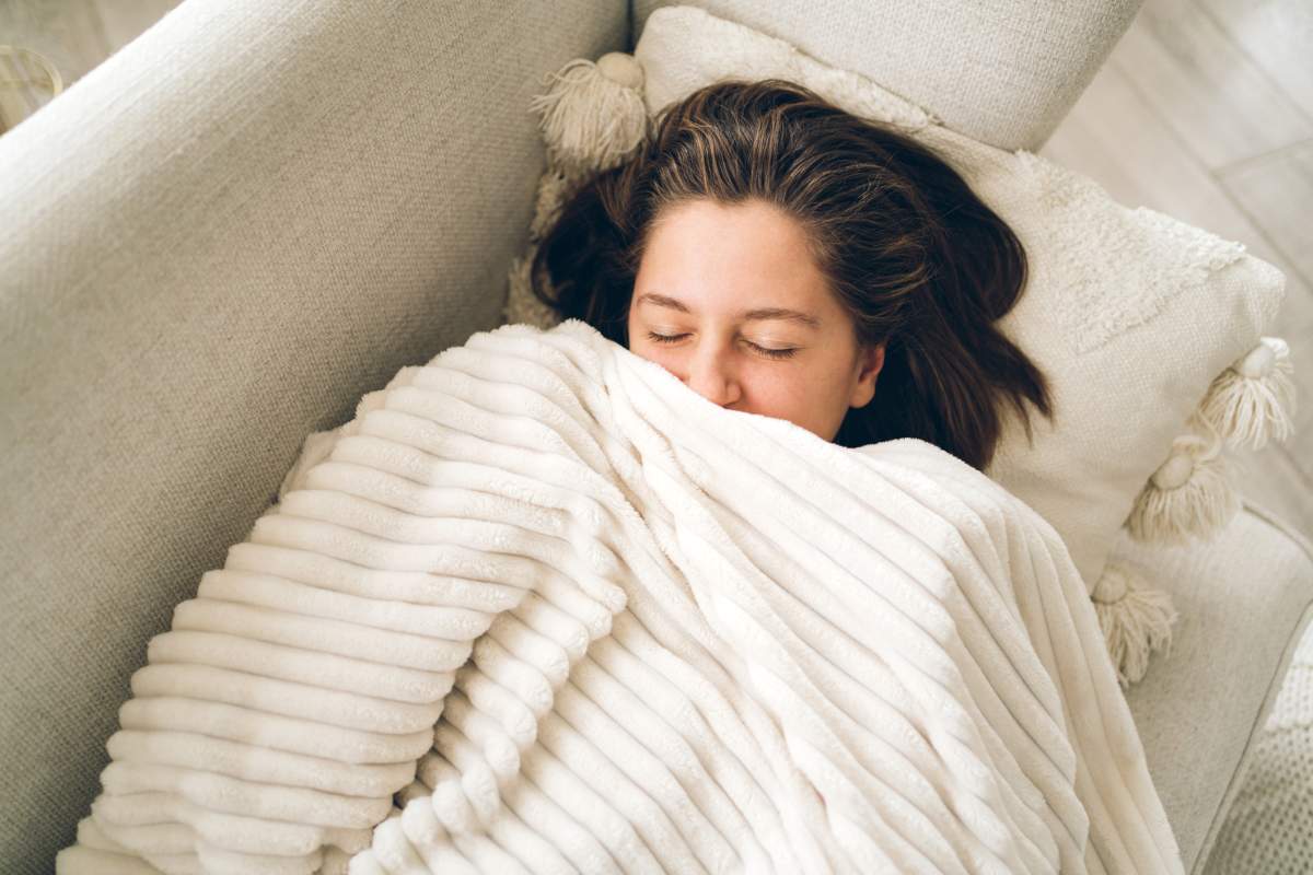 Young smiling woman lies on the couch and looks out from under the blanket. She has gray eyes and short brown hair. Photography from above