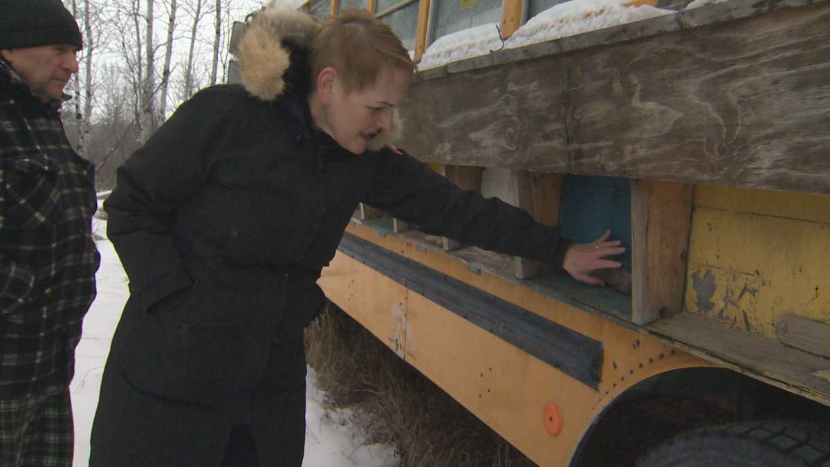 A woman in a black winter jackete touches the side of a modified school bus. An elderly man watches from her left.