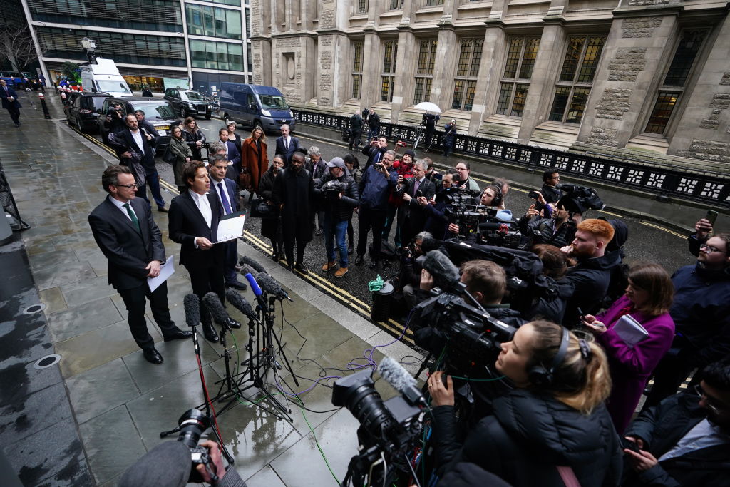Prince Harry's lawyer is seen speaking to a crowd of press on a rainy sidewalk.