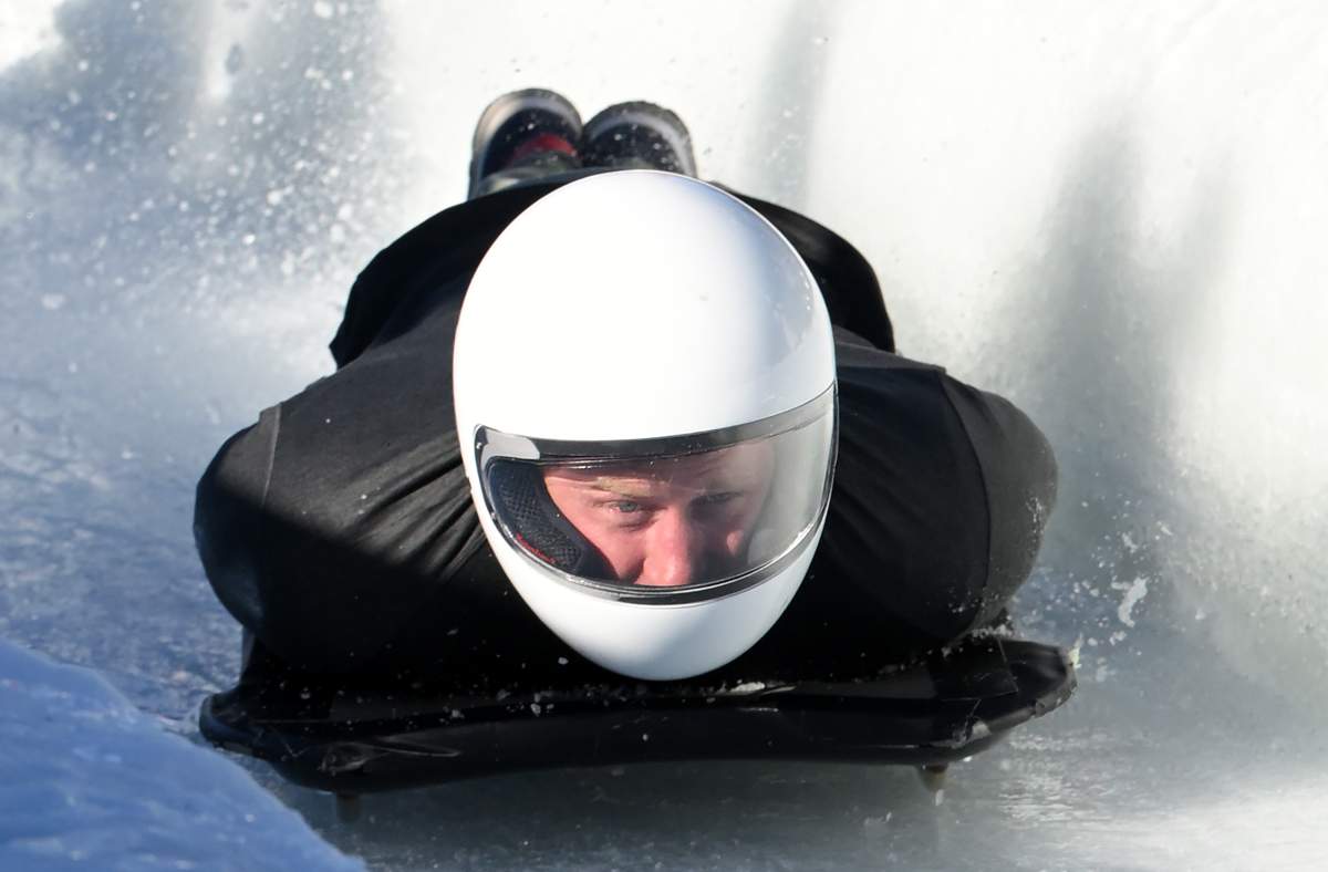 Prince Harry on a skeleton track. He is wearing black snow gear and a white helmet. He is sledding head-first.