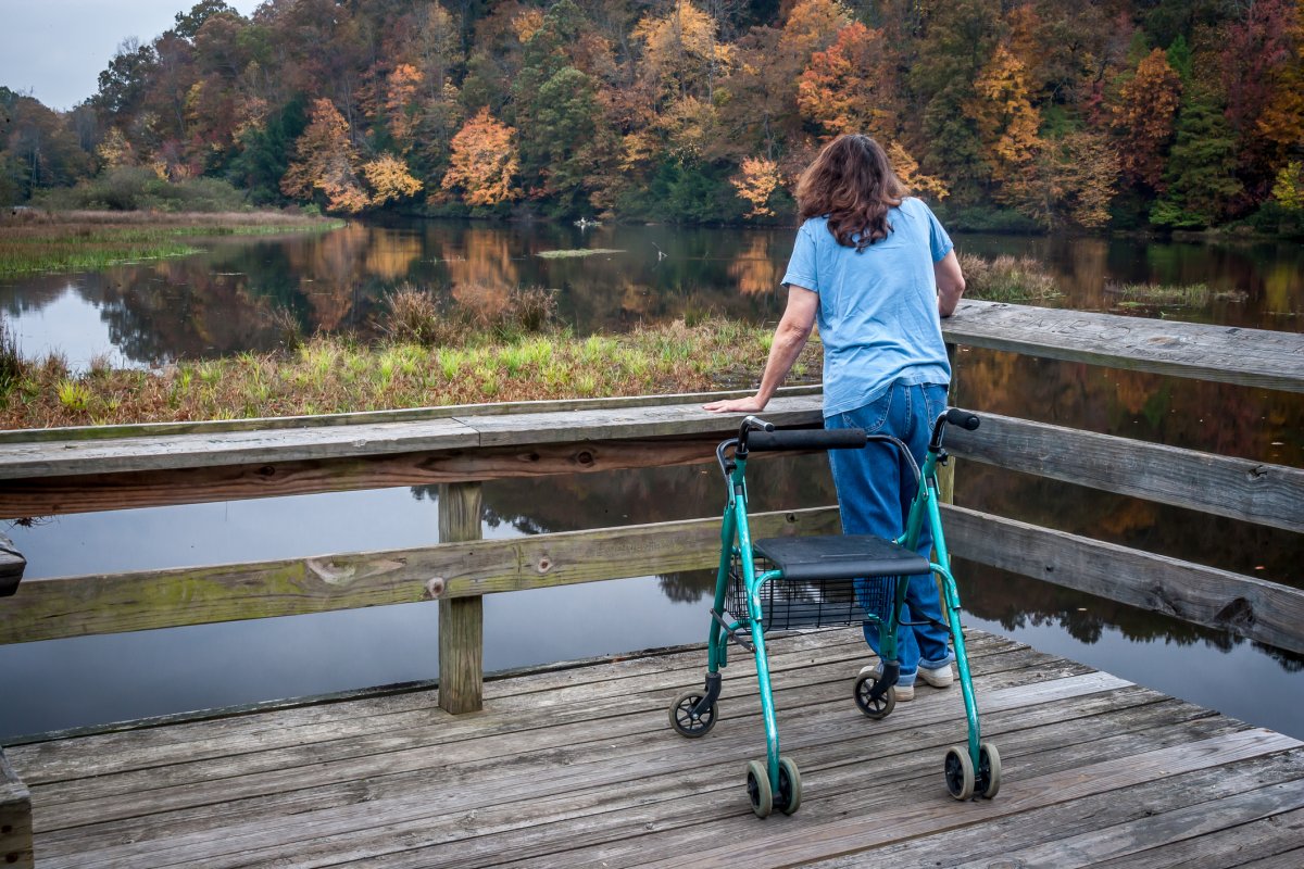 Handicapped lady with a walker stands on a dock looking out over calm lake at colorful fall foliage.