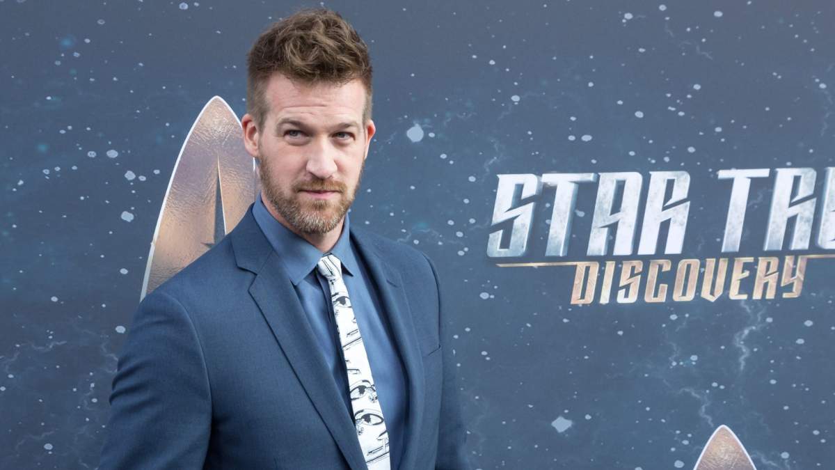 Kenneth Mitchell in a blue suit and white tie. He is on a red carpet in front of a Star Trek: Discovery banner.
