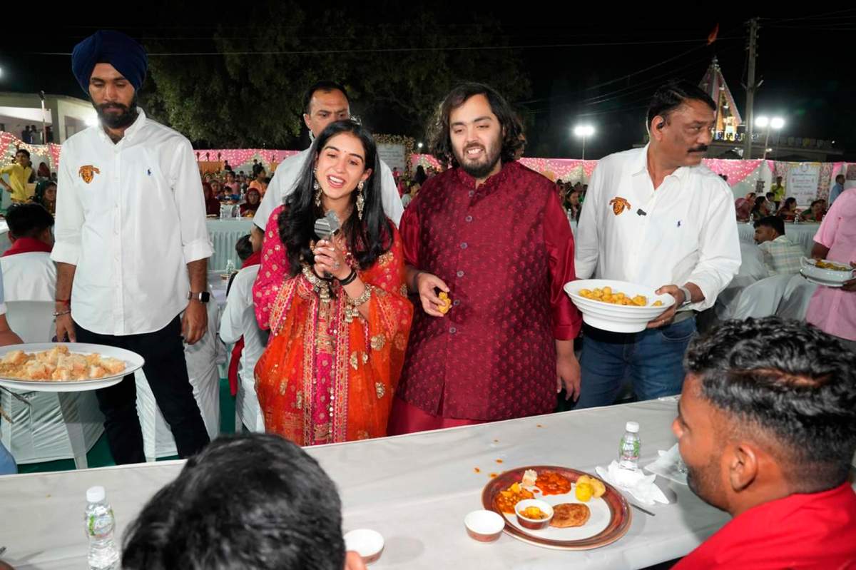 Anant Ambani and Radhika Merchant serve food and speak to locals. Radhika is holding a microphone. They are both wearing red,