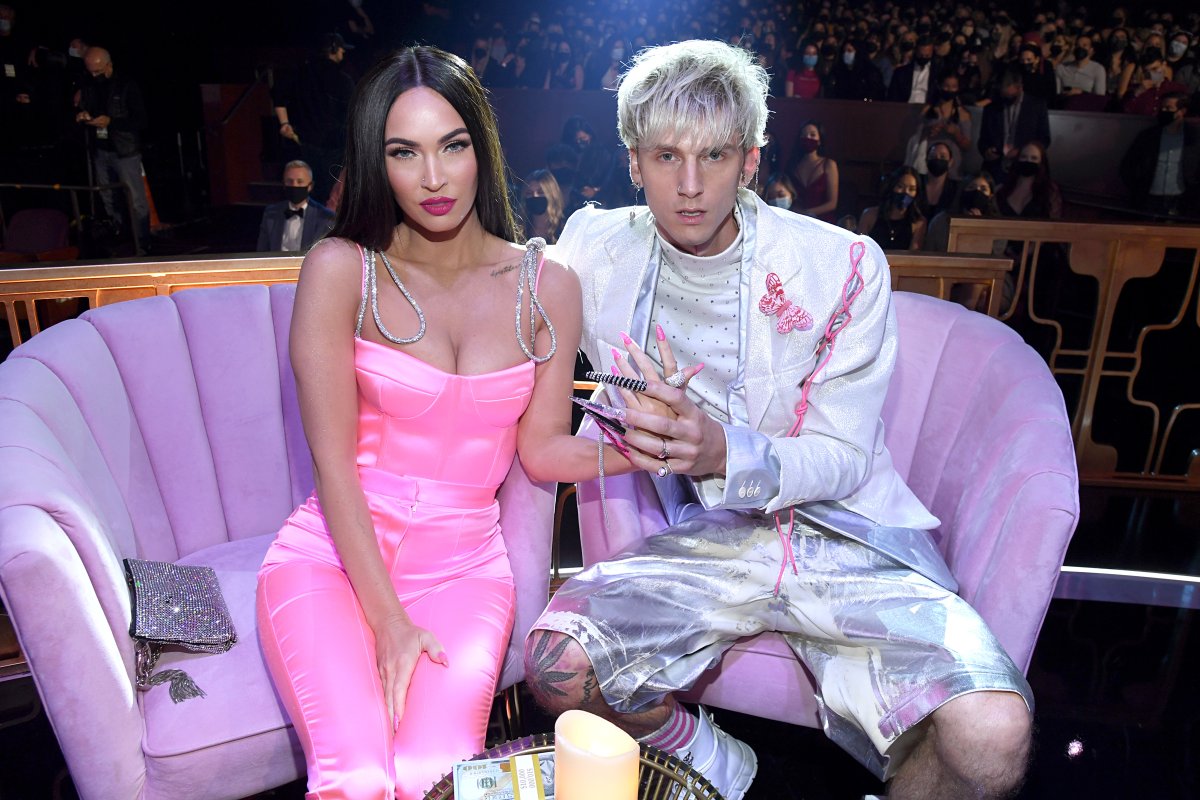 Megan Fox in a pink, sleeveless pantsuit. She is sitting next to Machine Gun Kelly, who is wearing silver shorts and a white suit jacket. They are holding hands.