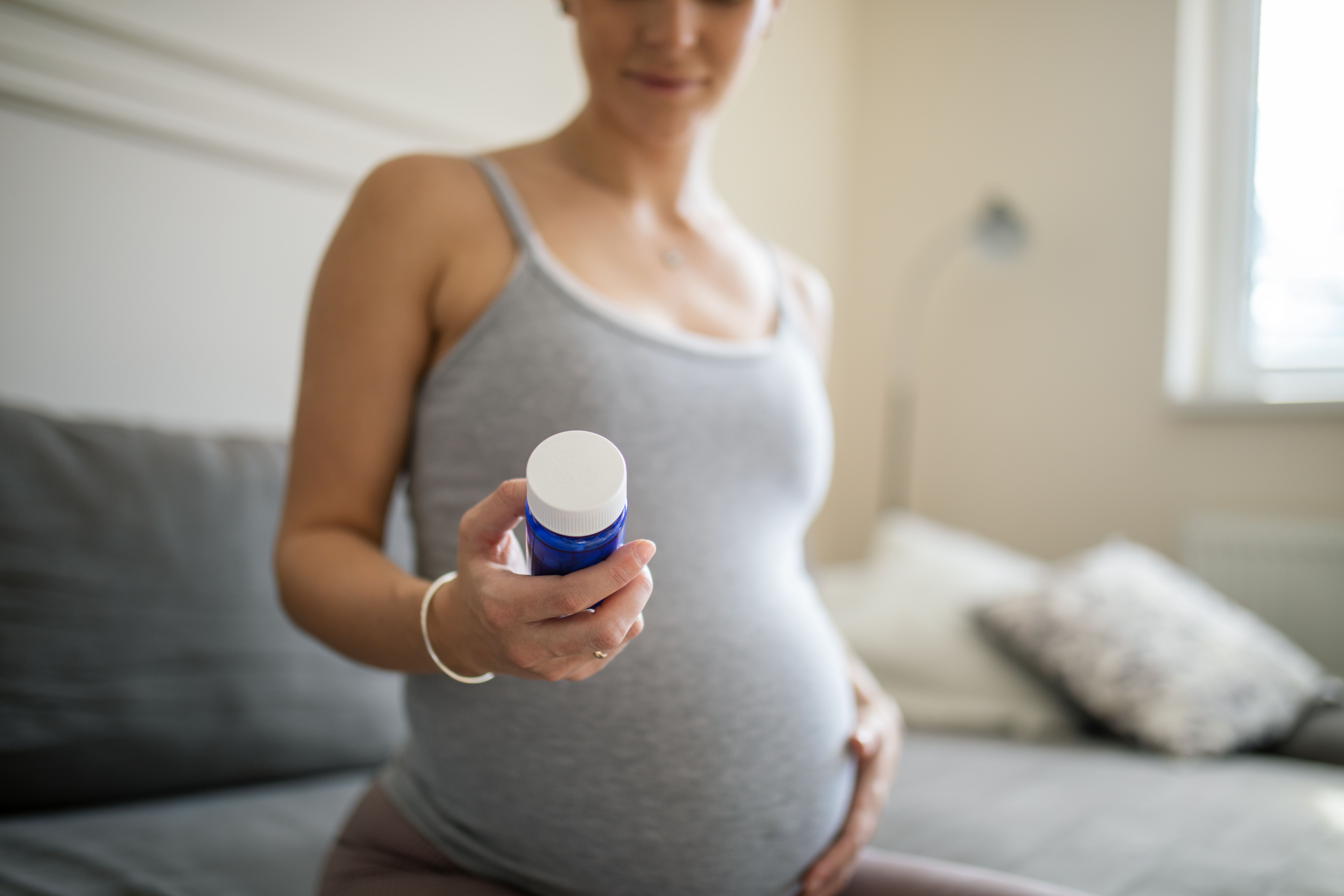 Beautiful young pregnant woman sitting on sofa in her living room and holding medical bottle with medicine pills or nutrition supplements and reading the label