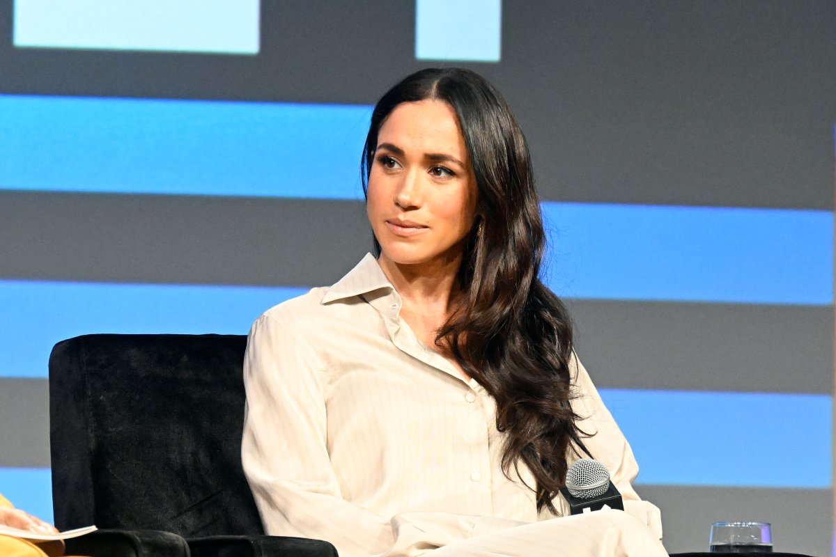 Meghan Markle. She is sitting, wearing a white button-up shirt. Her long hair is down and tucked to one side.