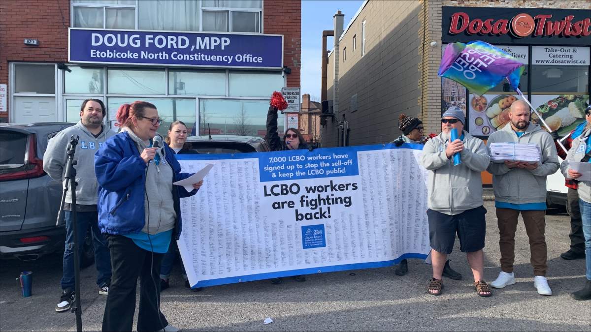 People holding a large banner in the background in front of Premier Doug Ford's constituency office while a woman holds a microphone in the foreground.