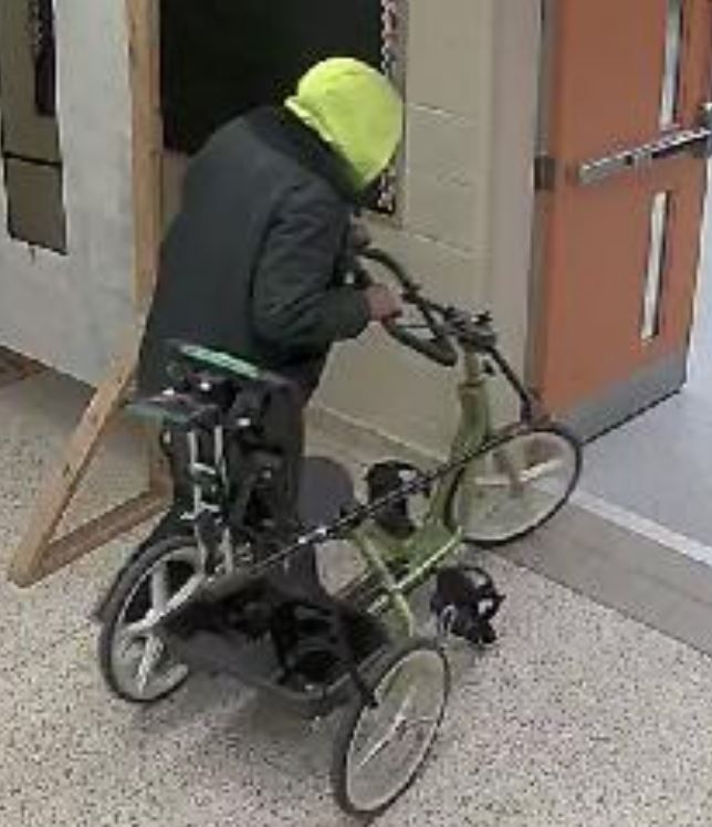 A surveillance image of a man in a hoodie wheeling an adaptive tricycle out of a building.