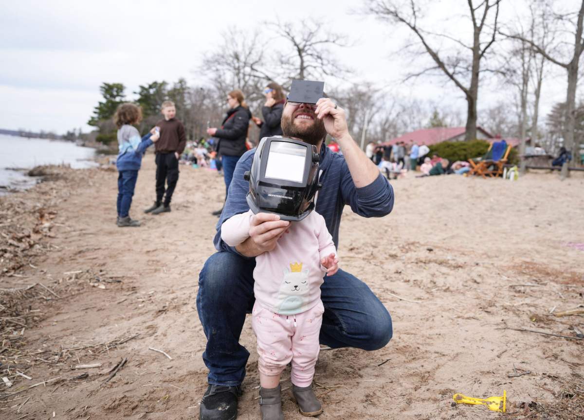 Jason Epstein and his two year old daughter Scarlett Epstein use a welder’s mask to watch the progress of the total solar eclipse at Verona Beach, N.Y., Monday, April 8, 2024. THE CANADIAN PRESS/Christinne Muschi