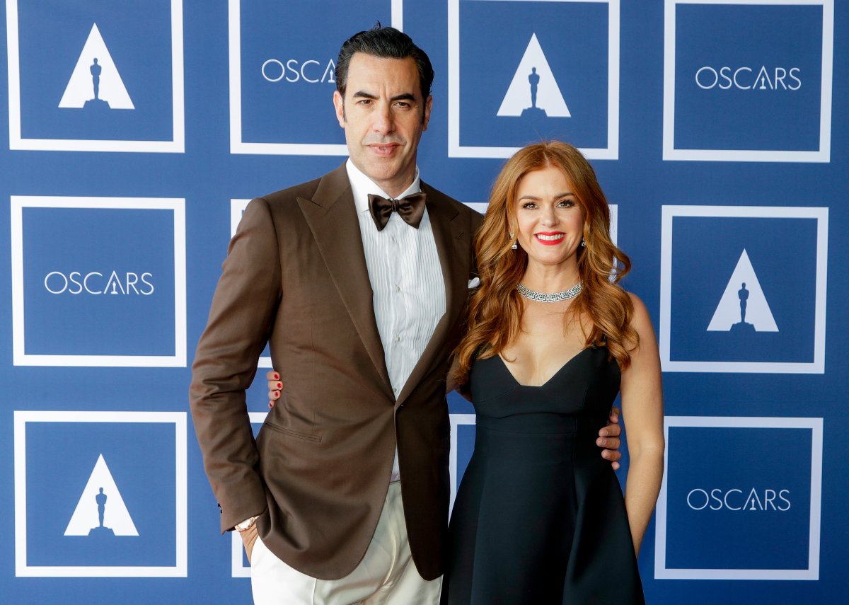 Sacha Baron Cohen and Isla Fisher on a red carpet. Baron Cohen, wearing a brown suit jacket and bowtie, has his arm around Fisher's waist. She is wearing a black dress.