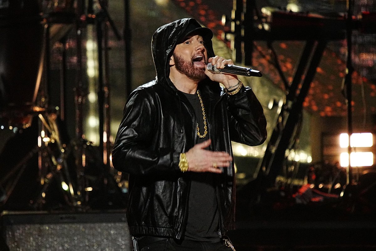 Eminem rapping on stage. He is wearing a black jacket with the hood up, a gold chain and a black tee.