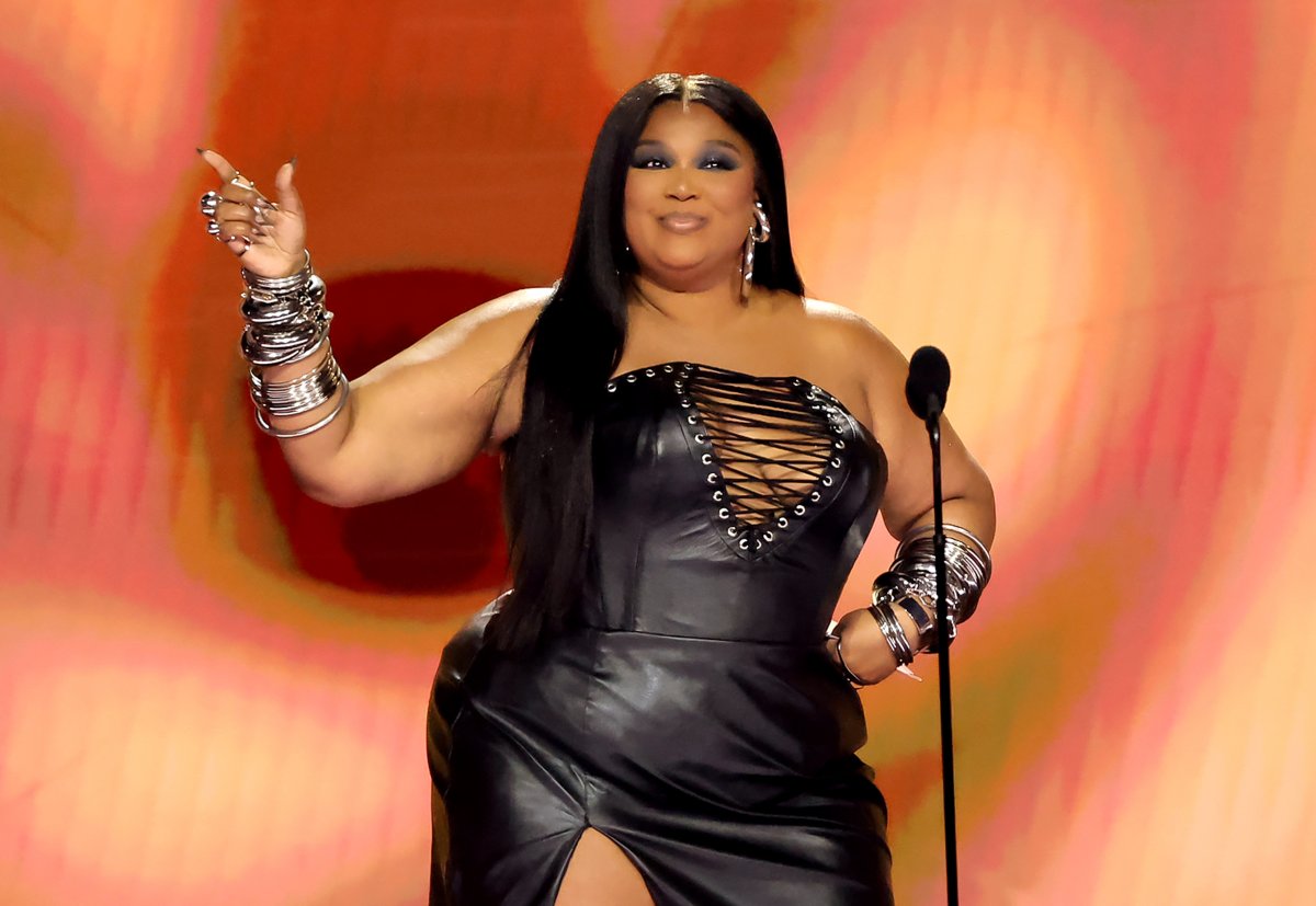 Lizzo on stage. She is wearing a black leather dress with an open cross section along the bust. Her hair is long and straight.
