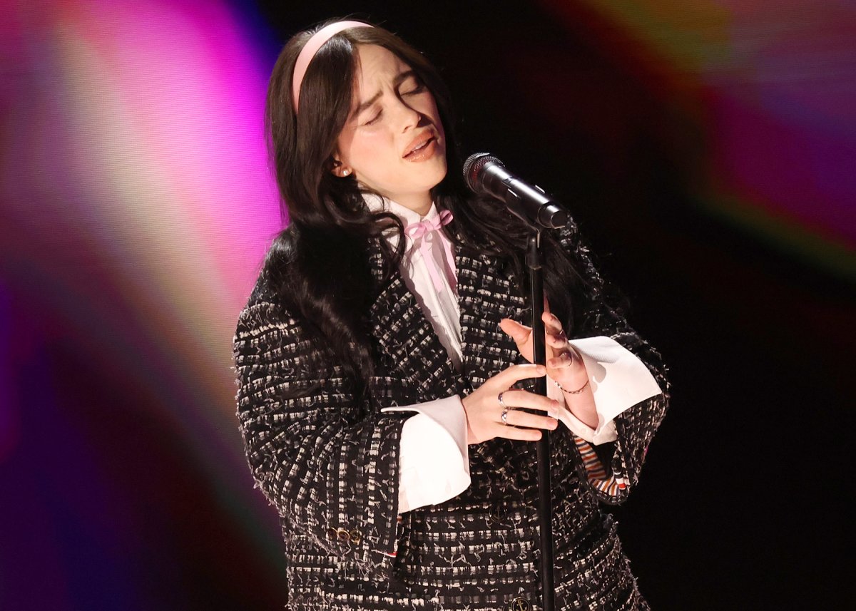 Billie Eilish singing on stage. She is wearing an oversized, jacket-blazer and a pink headband.