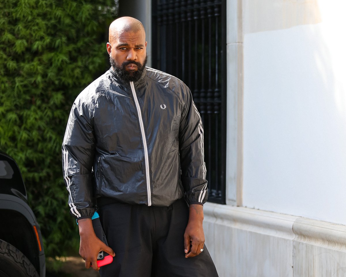 Kanye West (now Ye) outside. He is wearing a leather-type jacket and dark trousers. He is holding a cellphone and a Nintendo Switch.