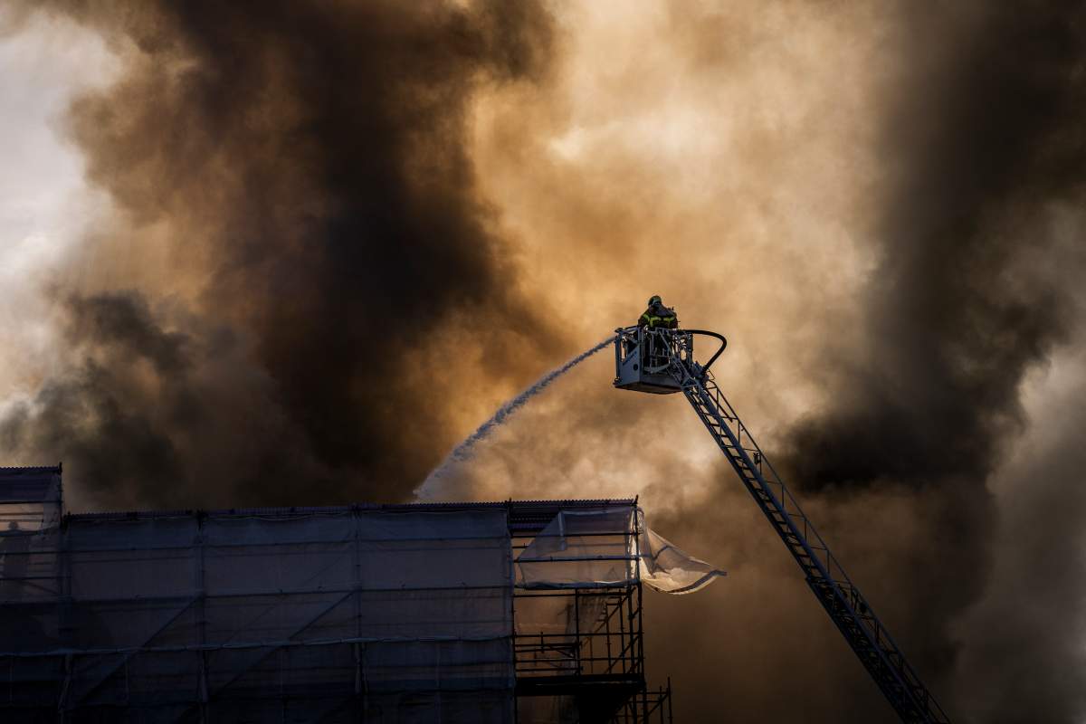A firefighter in an elevated, laddered bucket sprays water on a roof. Smoke plumes in the background.