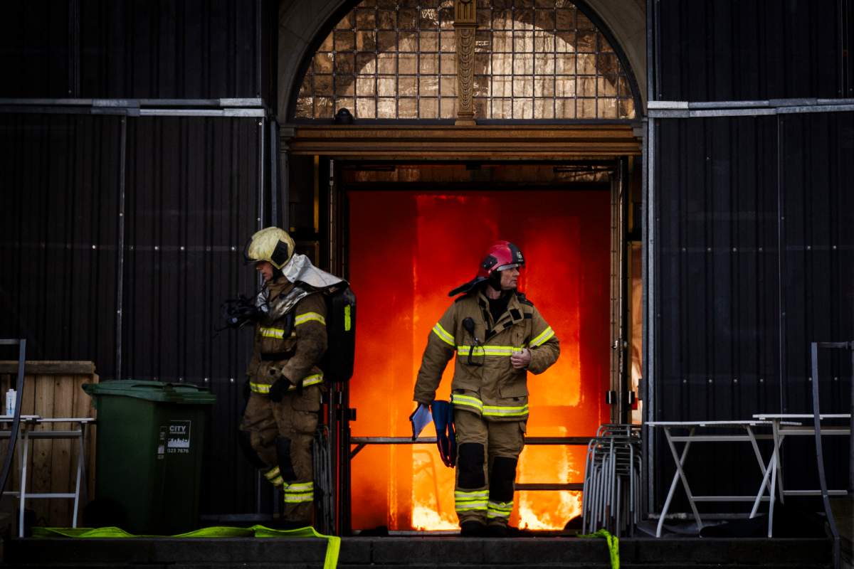 Two firefighters in a doorway that is backlit by orange flames.