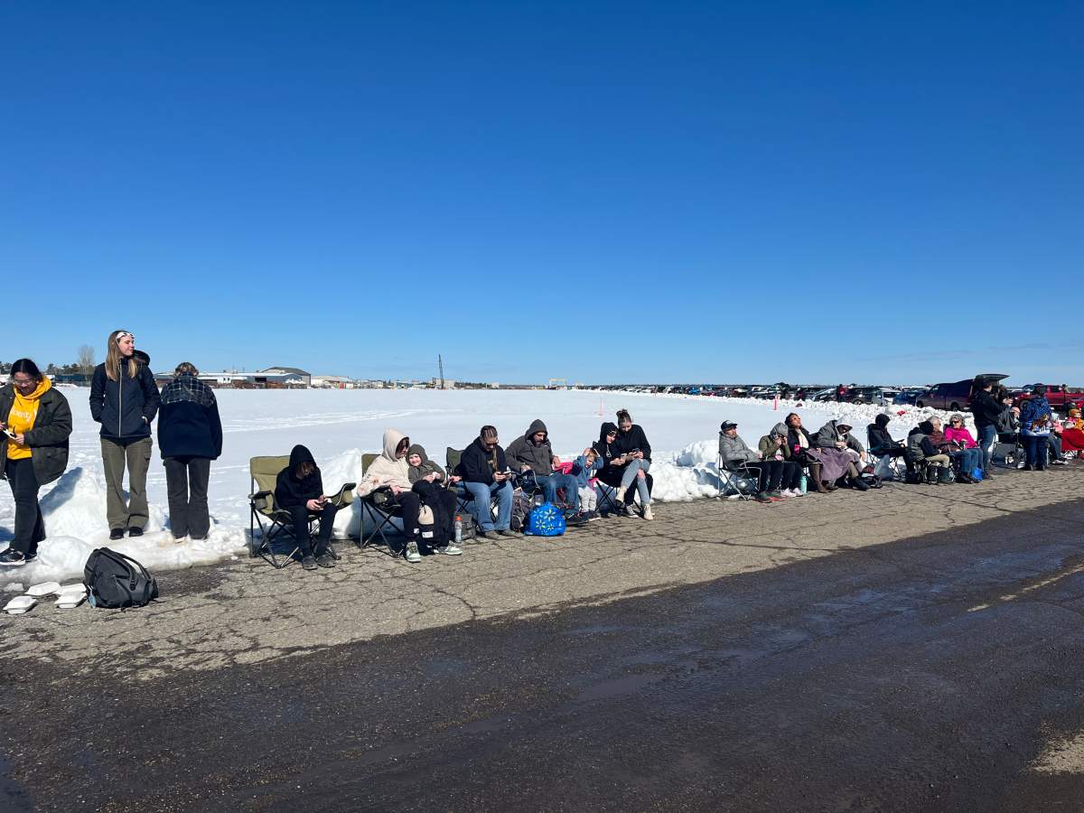 People standing along snow-covered ground by a road on a sunny day.