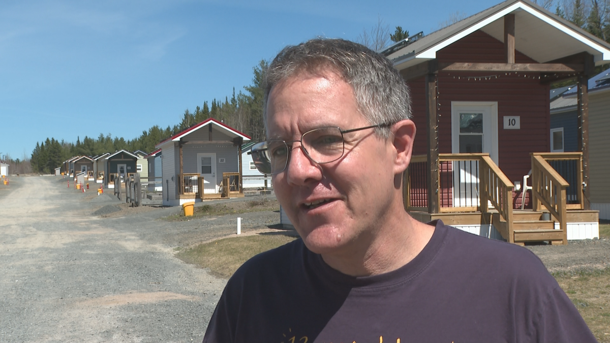 A man with a black shirt stands in front of rows of houses. Behind him to the right is a brown house with lights on it and the number 10.