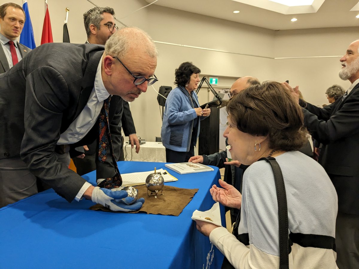 A man in a suit and glasses with gloves on holding a silver item leans over to speak with a woman.
