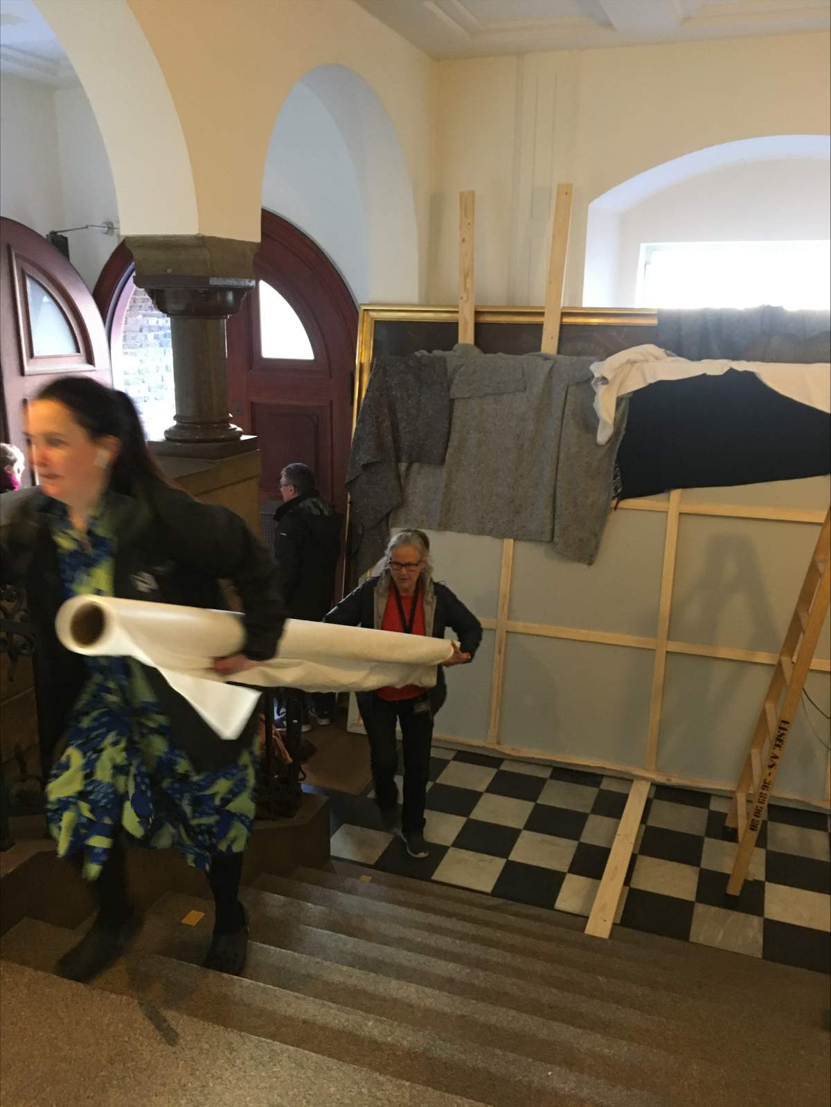 Two people carry a rolled canvas up stairs. Other canvases are propped up in the back.