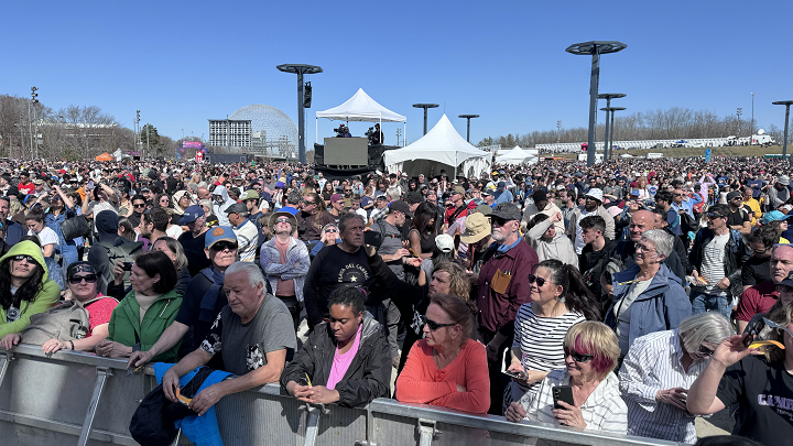 A crowd of people at a park on a sunny day standing behind a barricade.