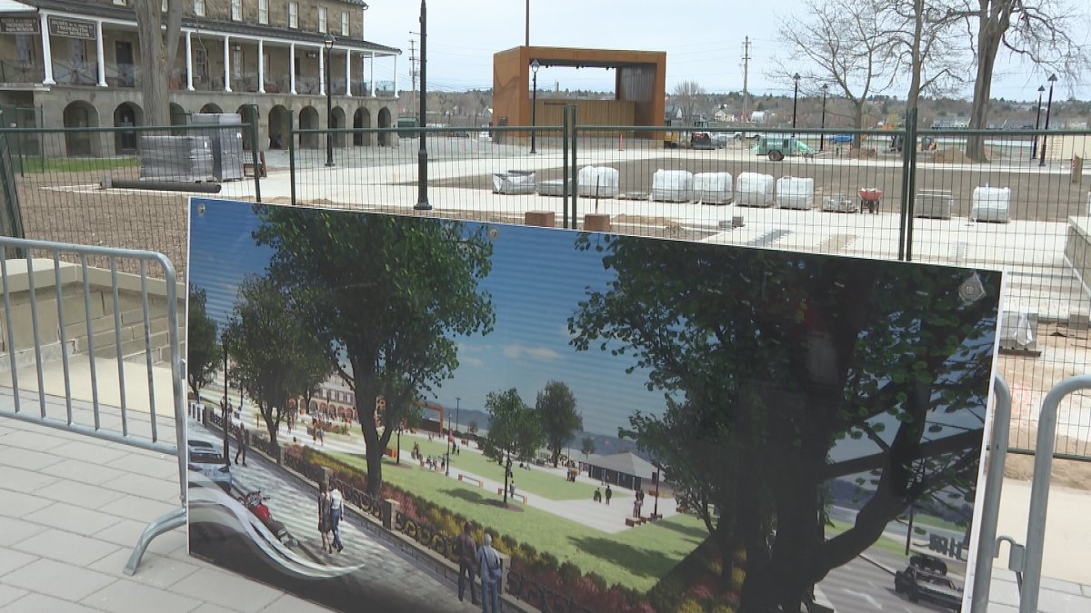 A sign showing a green space with people walking in and around it stands in front of a fenced-off space. In the background is a brown square structure.