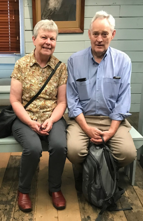 A man and a woman sit with their hands in their laps. They both are dressed casually, have short, white hair and are smiling.