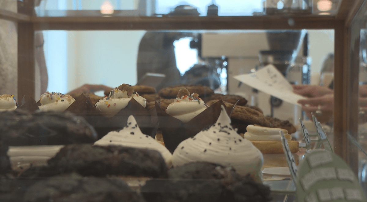 Rows of desserts line a clear display case. In the background, there are people's hands and a blurry figure stands at a coffee machine.