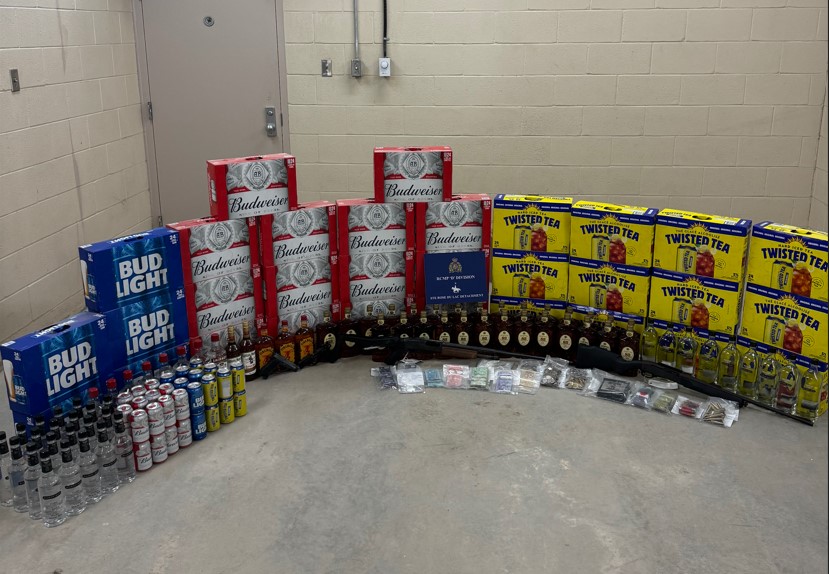 Several cases of beer and hard iced teas, as well as individual cans, bottles of liquor, a firearm, and bagged evidence are displayed against a white brick wall.