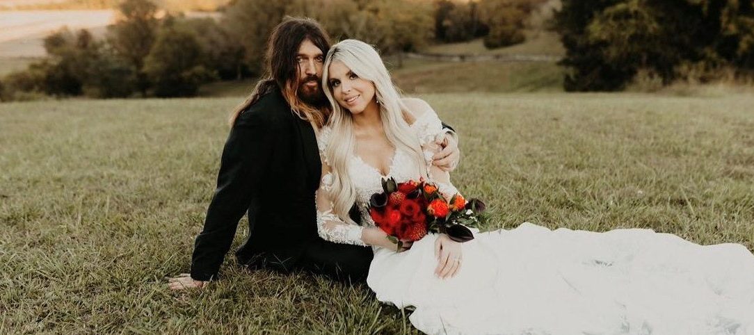 Billy Ray Cyrus and Firerose on their wedding day in October 2023. She is holding a red and orange bouquet and wearing a white dress. Cyris is wearing a black tux. They are laying on the grass.