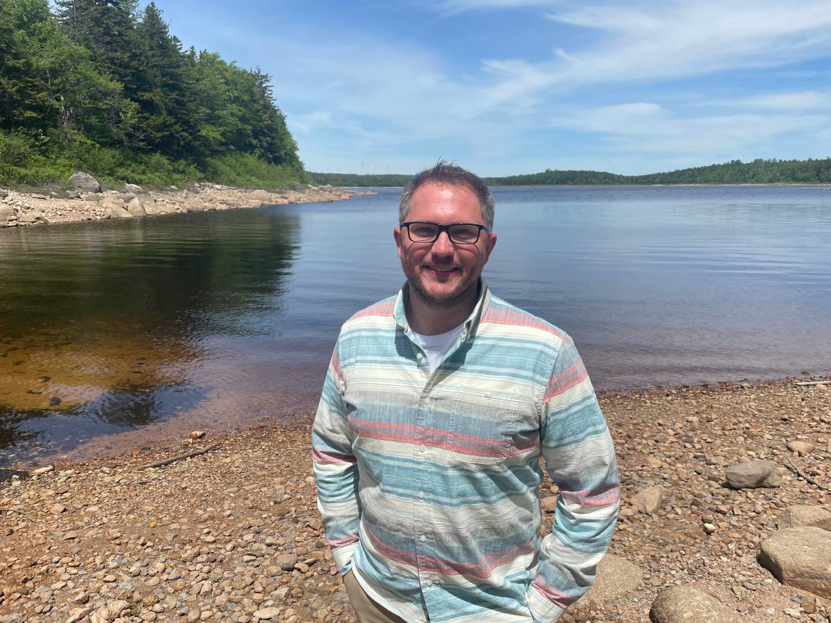 A man wearing a blue and grey striped shirt stands with his hands in his pants pocket on a sandy beach with a blue lake in the background, there are green trees on one side