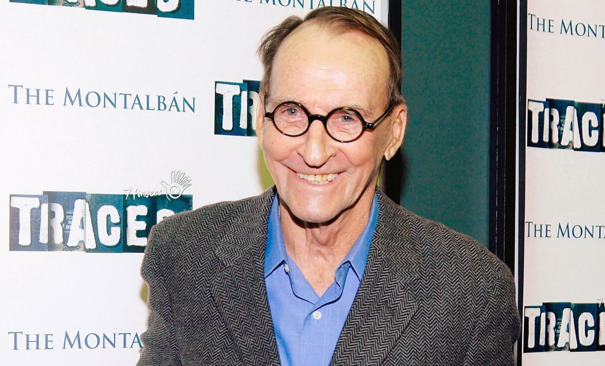 James Sikking in a grey suit jacket and blue shirt. He is wearing round glasses and smiling.