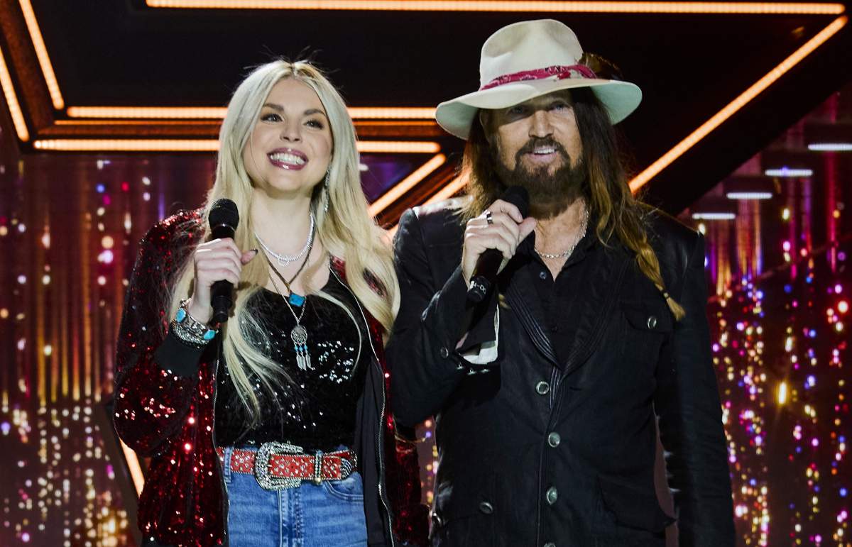 Firerose and Billy Ray Cyrus stand on a stage holding mics.