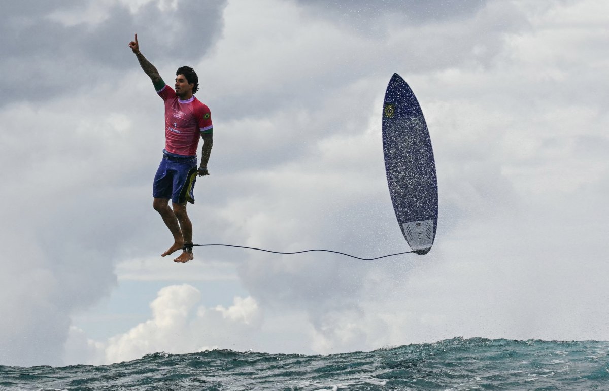 Gabriel Medina, mid-air next to his parallel surfboard. He is pointing his index finger at the sky.