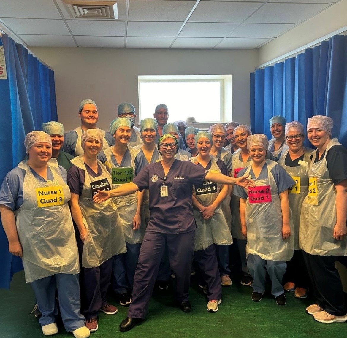 Nurses and doctors and midwives stand for a photo in their scrubs and caps. Many are wearing tape on their chest that notes an order, like "Nurse 1, Quad 3."