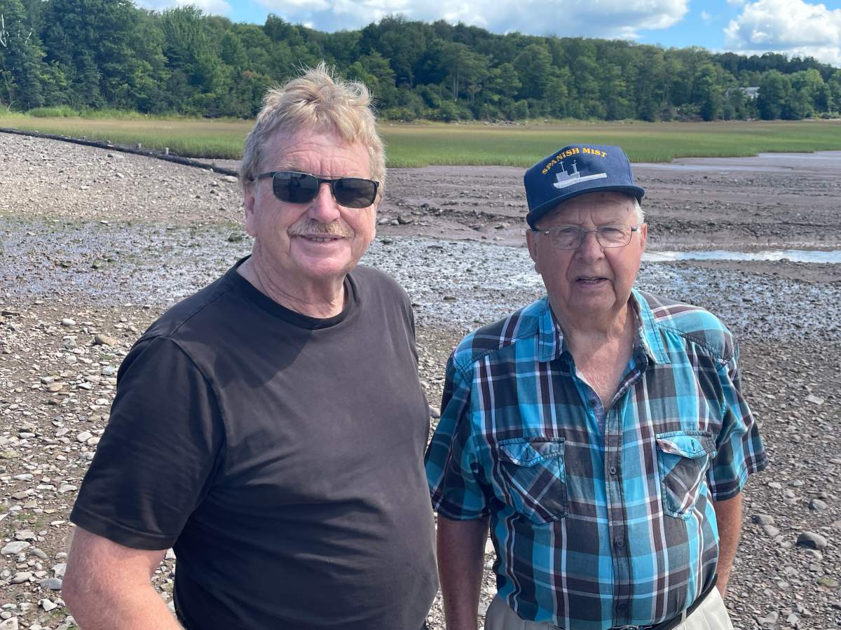 Two men stand next to each other. Once man is wearing sunglasses and a dark t-shirt. The other is a senior wearing a blue cap and blue plaid shirt.
