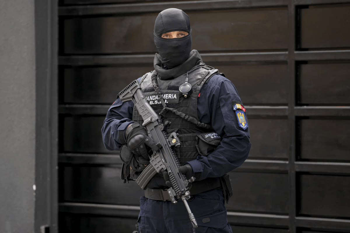 A masked officer in black clothing, a bulletproof vest and holding a rifle.